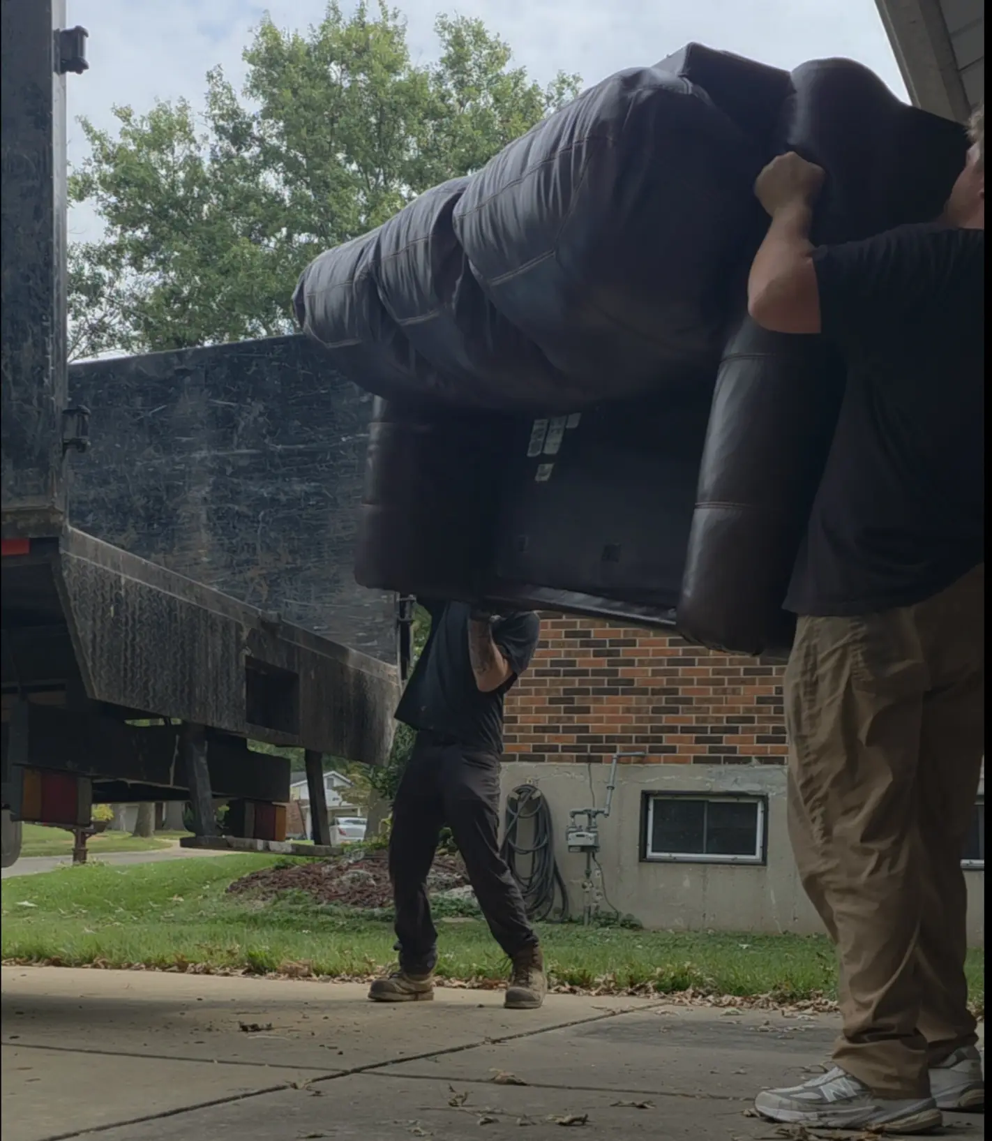Two crew members carrying a brown leather couch overhead down the driveway to the truck