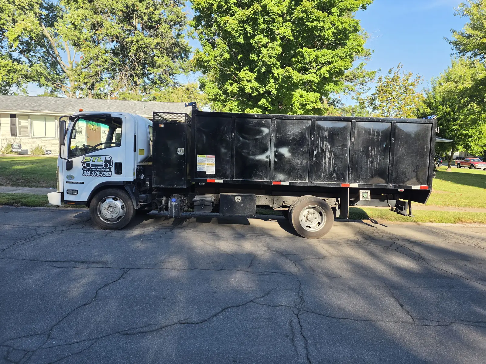 White box truck with a high-sided black dump bed, company logo and phone number on the door, parked on a residential street